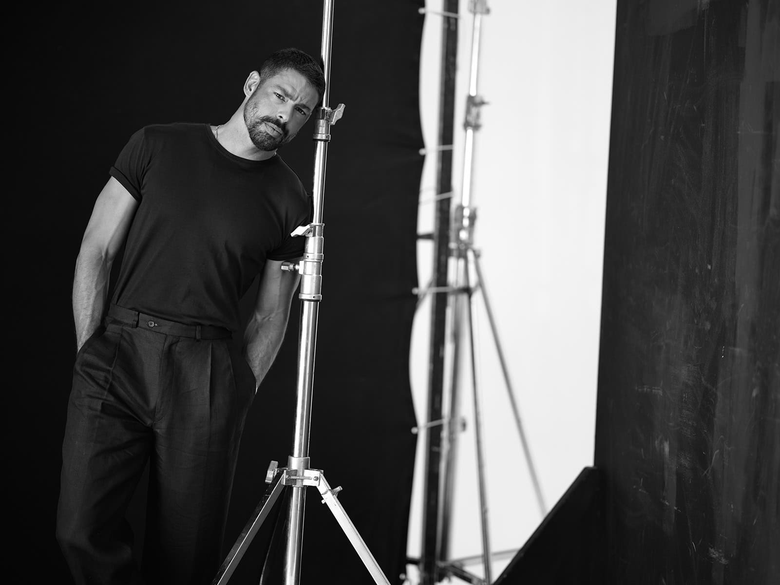 Retrato em estúdio de um homem com expressão séria, posando ao lado de equipamento de iluminação em ambiente profissional de fotografia.