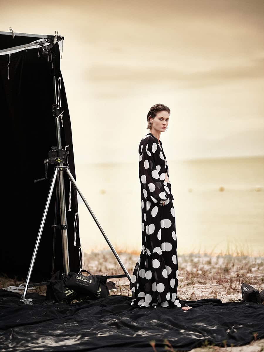 Foto de uma modelo posando ao ar livre durante uma sessão de fotos, usando vestido longo de estampa de bolinhas pretas e brancas, com iluminação profissional e fundo de praia ao entardecer, destaque para produção fotográfica de alta qualidade e moda.