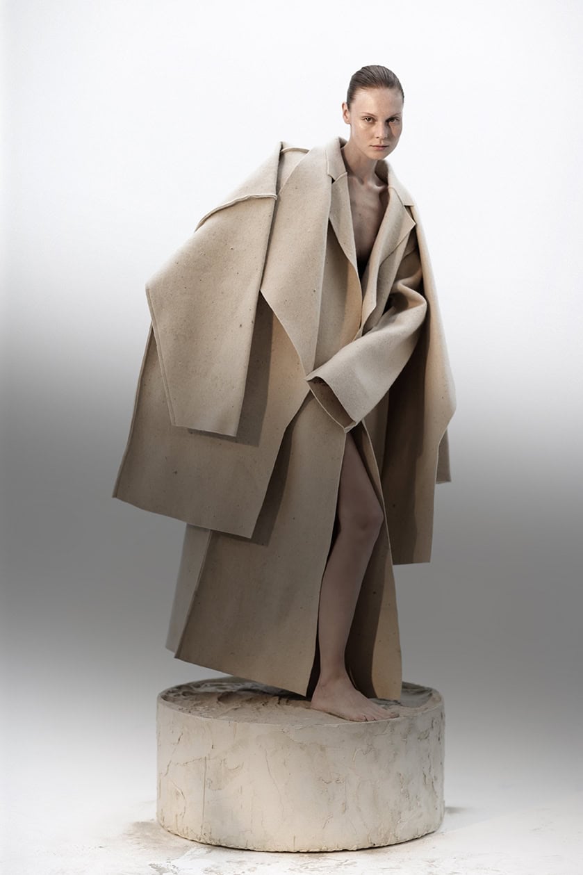 Imagem de uma modelo usando um porte de casaco bege, em um ensaio de moda estilizado e minimalista, com fundo branco e pedestal decorativo, destacando elegância e sofisticação em produção fashion.