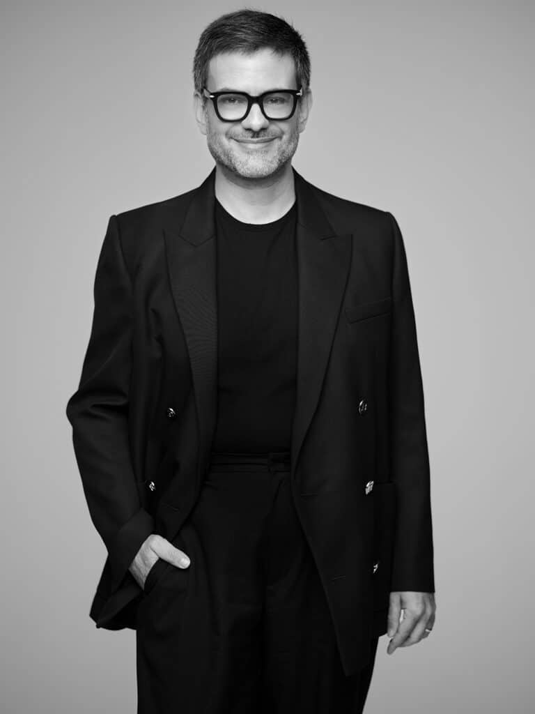 Fotografia de um homem elegante de óculos escuros, sorriso confiante, vestindo terno preto, imagem em preto e branco, estilo profissional, ideal para branding e conteúdo corporativo, foco em confiança e sofisticação.