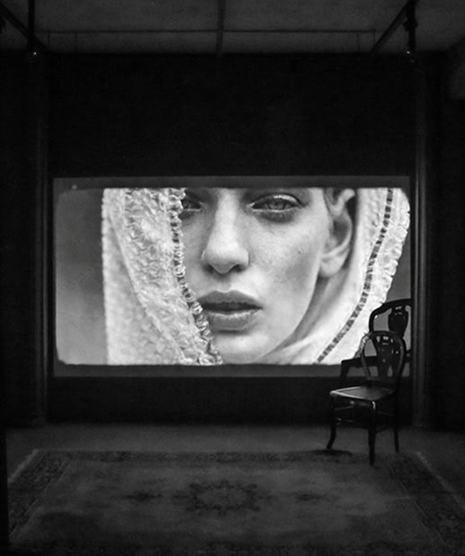 Fotografia em preto e branco de um rosto de mulher projetado em uma tela grande, com uma cadeira ao lado, criando um ambiente artístico e moderno para ensaios fotográficos.